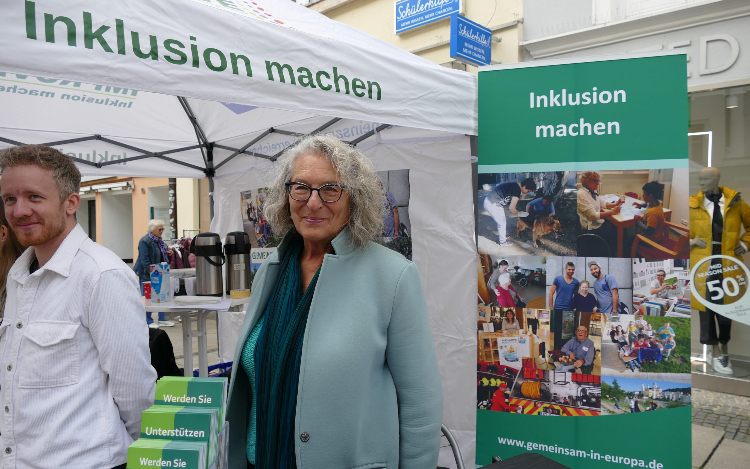 Eine Frau und ein Mann stehen vor einem Pavillon und einem Poster. Auf beiden steht: Inklusion machen.