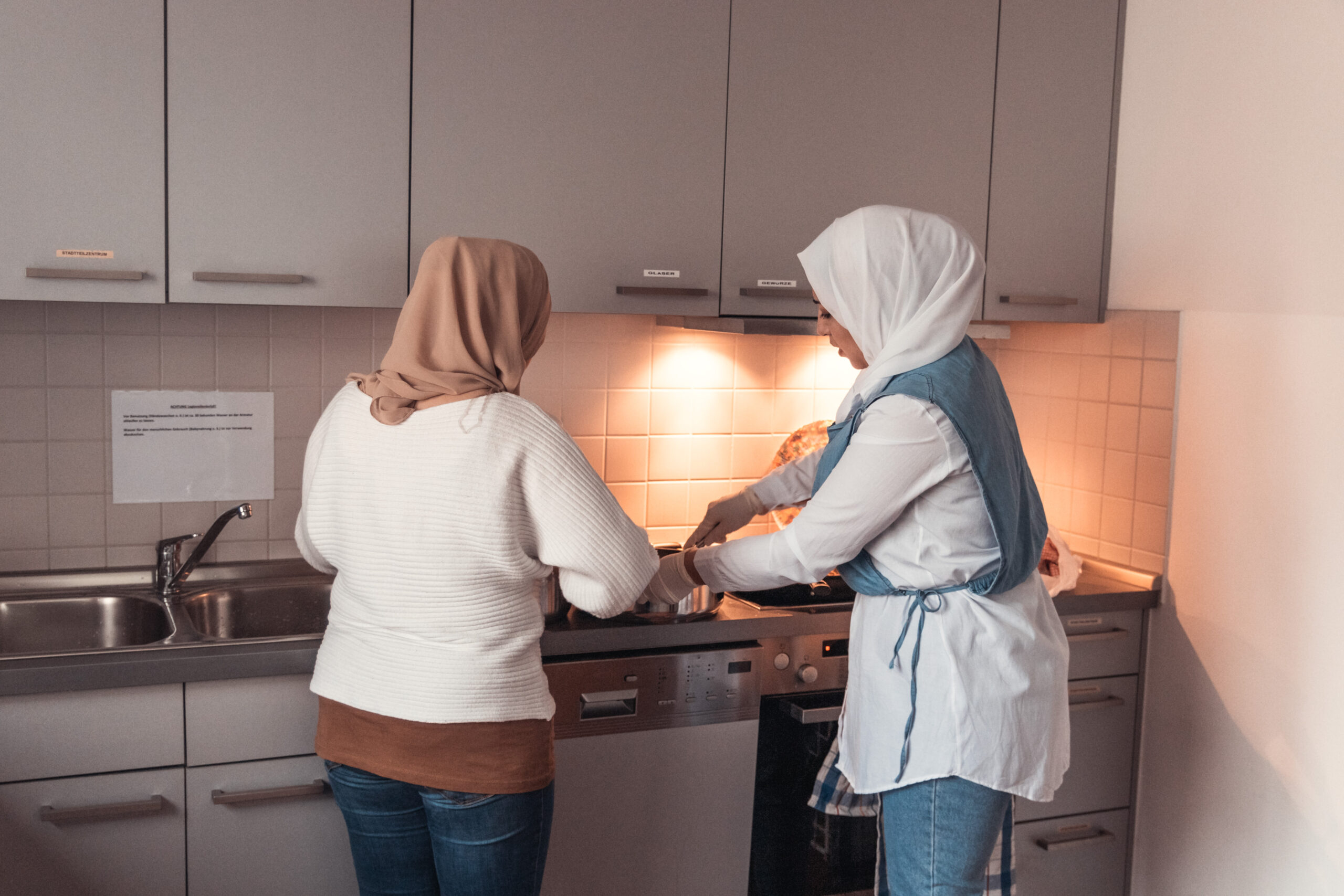 zwei Frauen mit Kopftuch stehen mit dem Rücken zur Kamera und kochen.