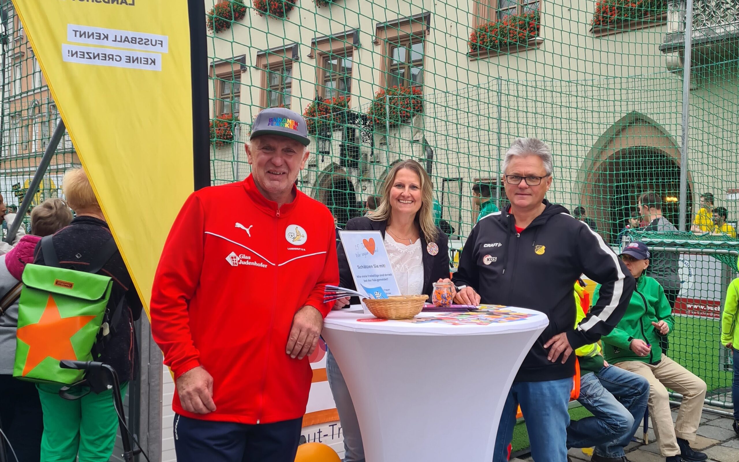 2 Männer und eine Frau stehen an einem Stehtisch. Im Hintergrund sind mehrere Personen, ein Fußballnetz und ein Hauswand zu sehen.