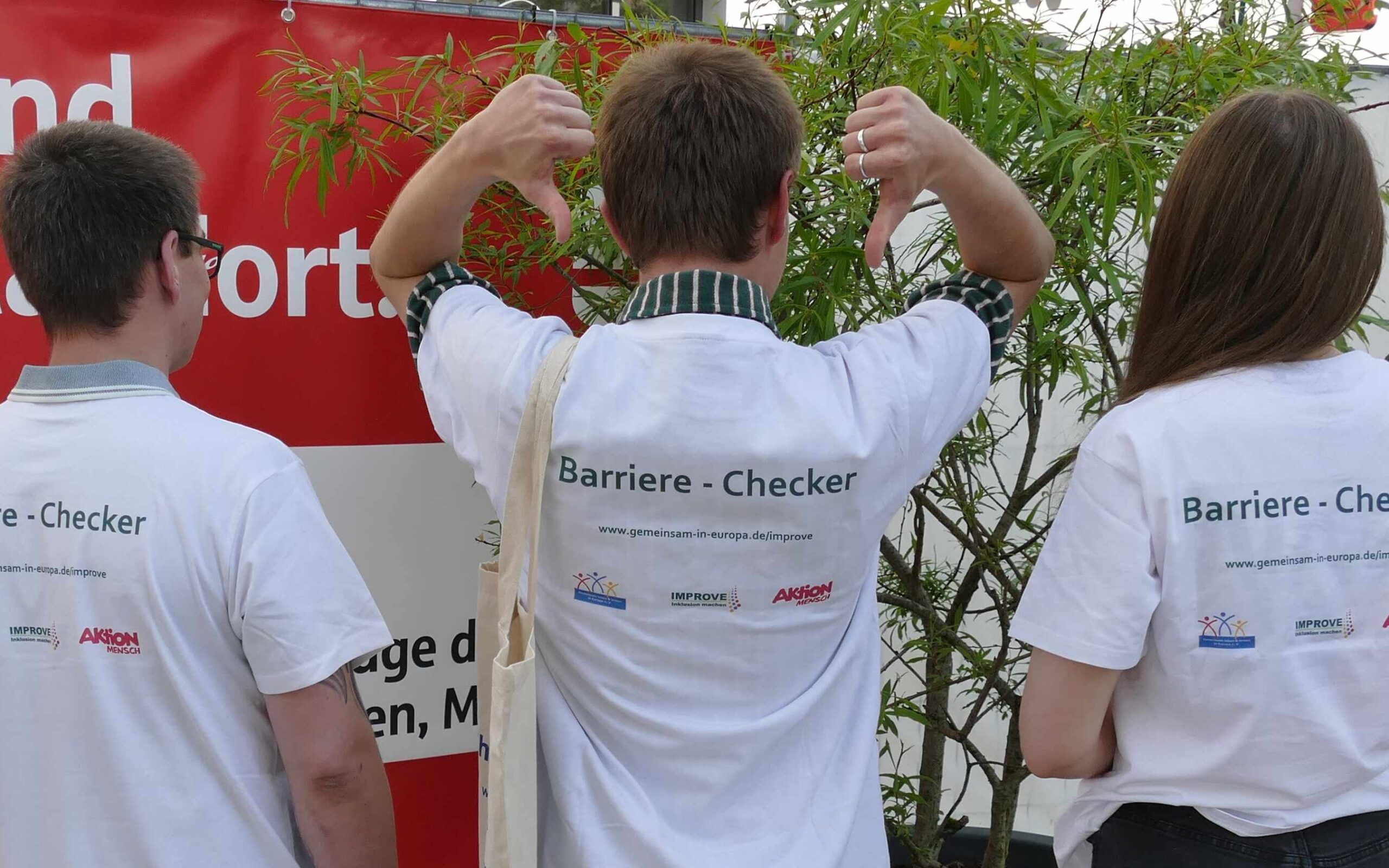 Man sieht drei Personen, eine Frau und 2 Männer, die mit dem Rücken zur Kamera stehen. Sie tragen weiße T-Shirts mit schwarzer Schrift 