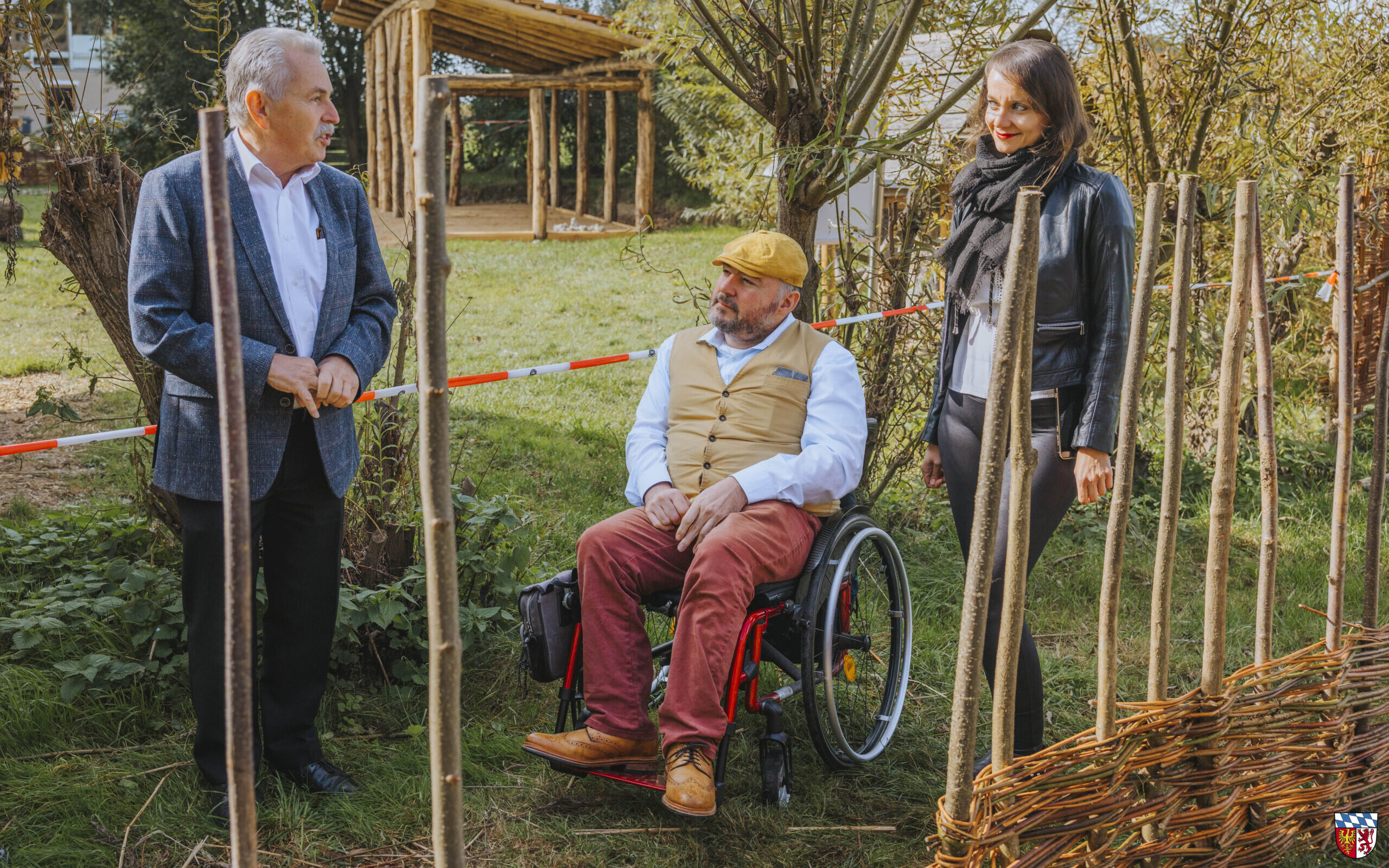 Eine Frau und 2 Männer, einer davon im Rollstuhl, befinden sich draußen in einem Garten. Im Vordergrund kann man einen halbfertigen Weidezaun sehen.