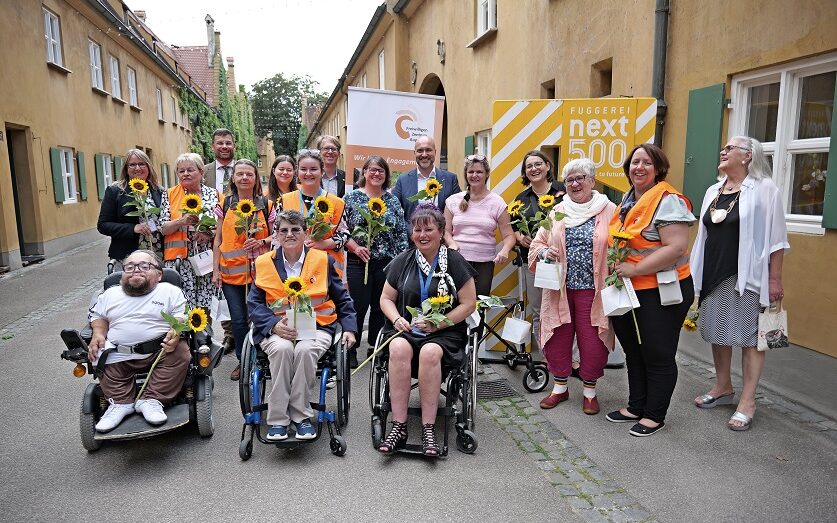Eine große Gruppe von Menschen,13 Frauen und 4 Männer, darunter drei Rollstuhlfahrer, stehen in der Fuggerei Augsburg. Alle haben eine Sonnenblume in der Hand.