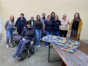 Ein Gruppenbild von 11 Personen, 3 Männer und 8 Frauen. Sie stehen vor einer Hauswand. Im Vordergrund ist ein Biertisch aufgebaut auf dem Teile einer Legorampe stehen. Neben dem Tisch ist einer der drei Männer, er sitzt im Rollstuhl